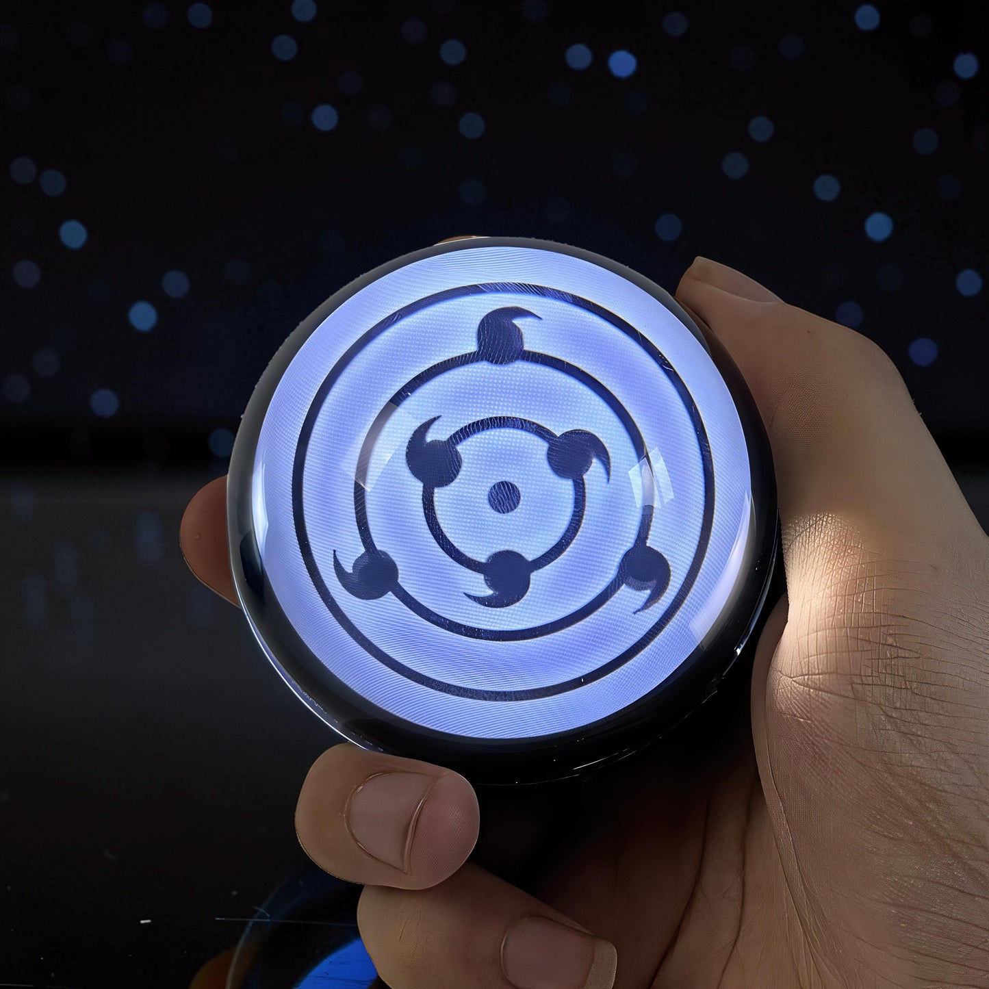The Curious Mind Sharingan Crystal Ball Decorative Ornament, a blue-lit crystal resin sphere with a black Sharingan eye design, held in a hand.