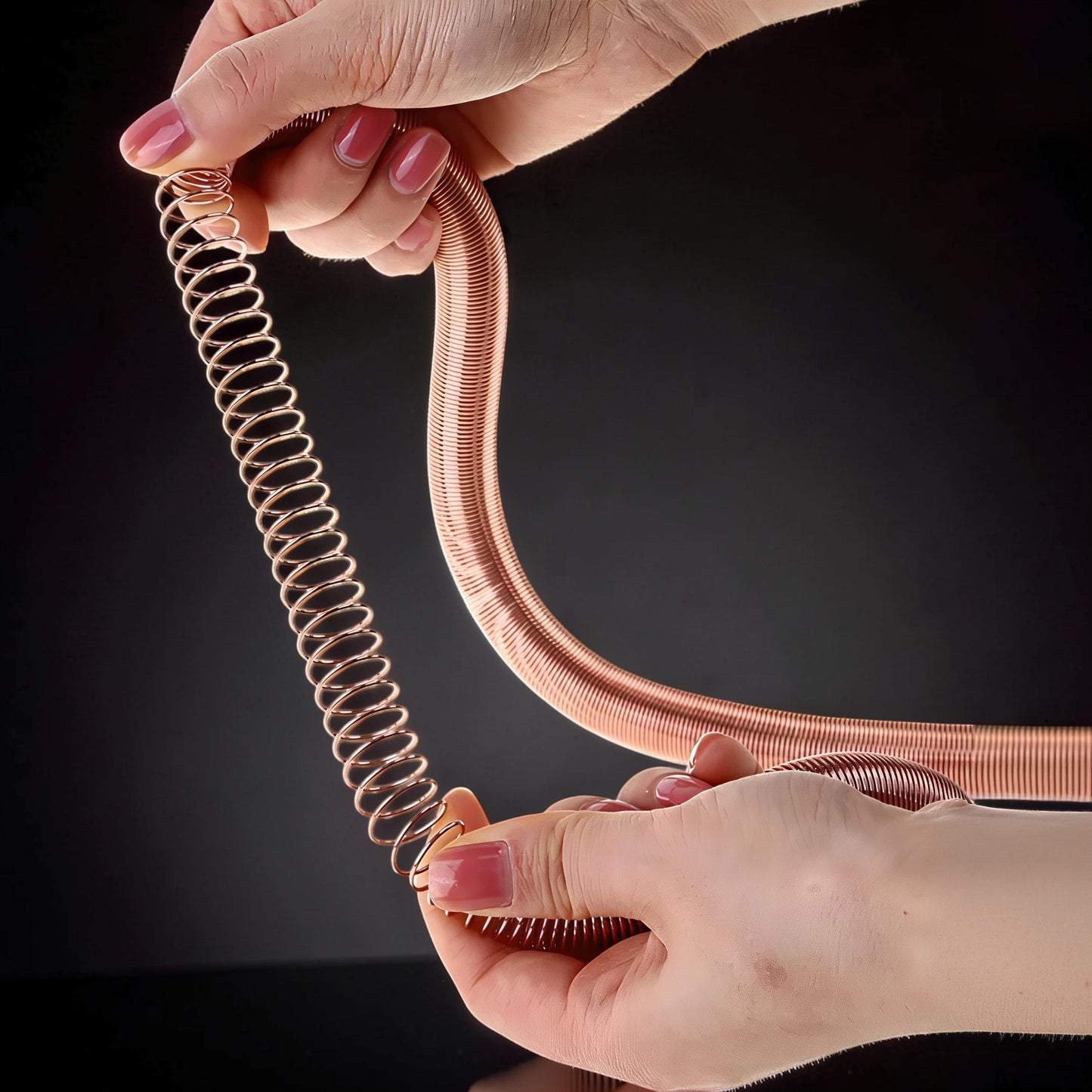 The Curious Mind Electromagnetic Train Puzzle Toy, a copper-colored metal spring coil, is shown being manipulated by two hands.