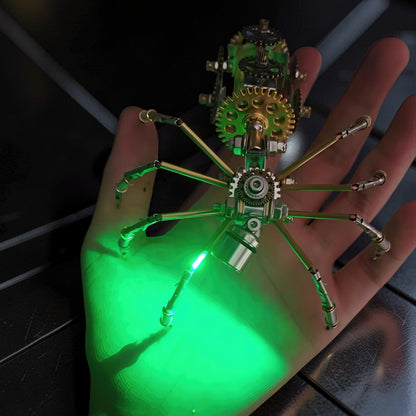 The Curious Mind Style E mechanical spider DIY kit, made of metal, with intricate gears and a green LED light, resting on a hand.