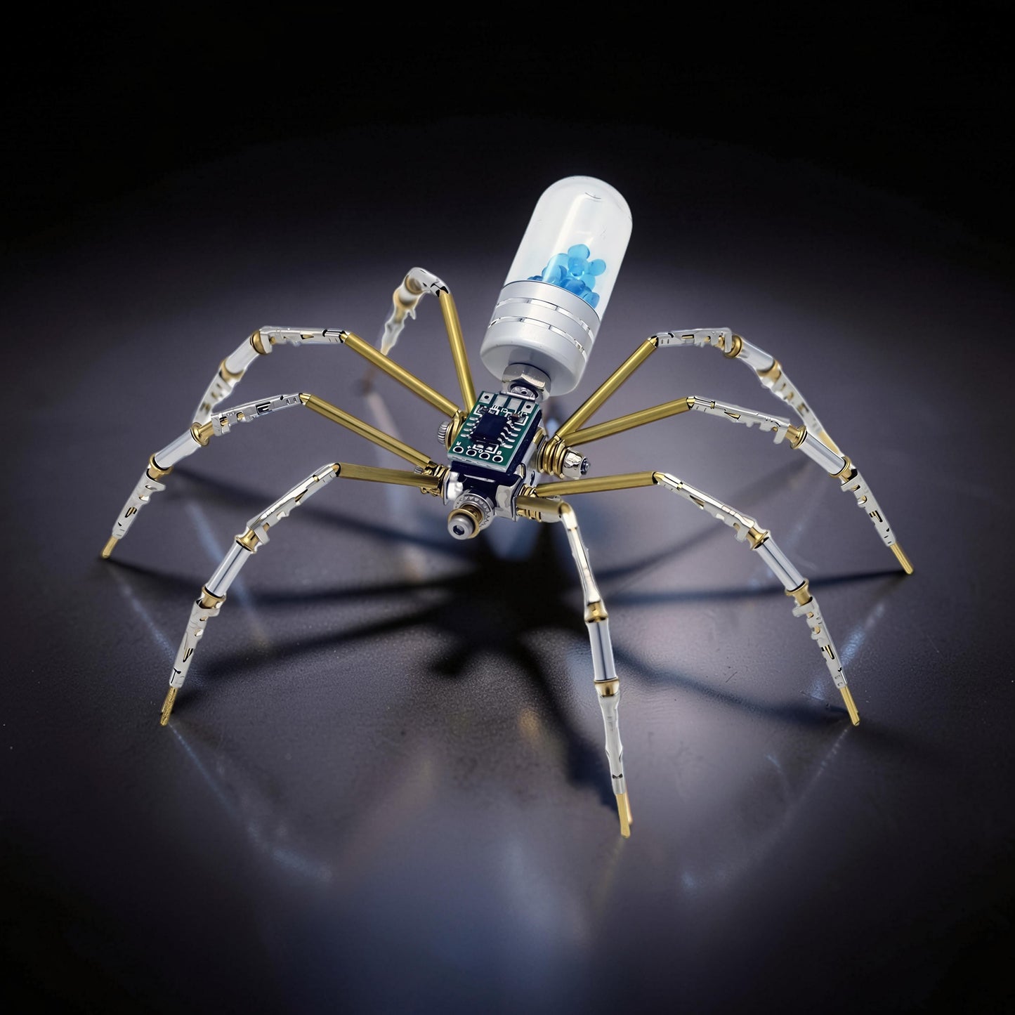 The Curious Mind Style B mechanical spider model kit, made of metal with brass accents and a clear capsule containing blue beads.
