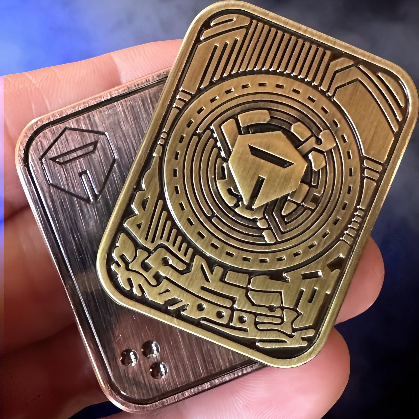The Curious Mind Three-Body Push Card Fidget in brass and bronze metal with intricate maze-like design and a central helmeted icon.