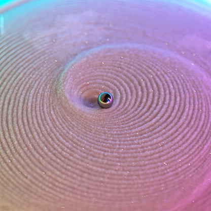The Curious Mind SANDSARA Mini Sand Painting Machine with a chrome ball creating concentric circles in fine sand, illuminated with pink and blue light.