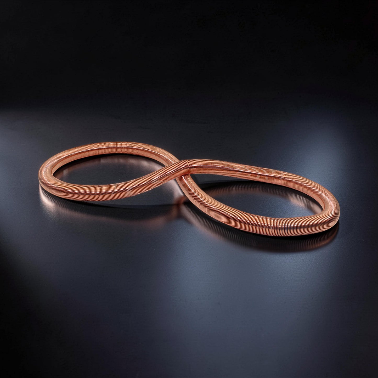 The Curious Mind Electromagnetic Train Puzzle Toy, a copper-colored metal coil in an infinity shape, displayed on a dark reflective surface.