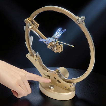 The Curious Mind's gold metal kinetic dragonfly model kit, a mechanical creature with blue wings, displayed on a stand with a large gear.