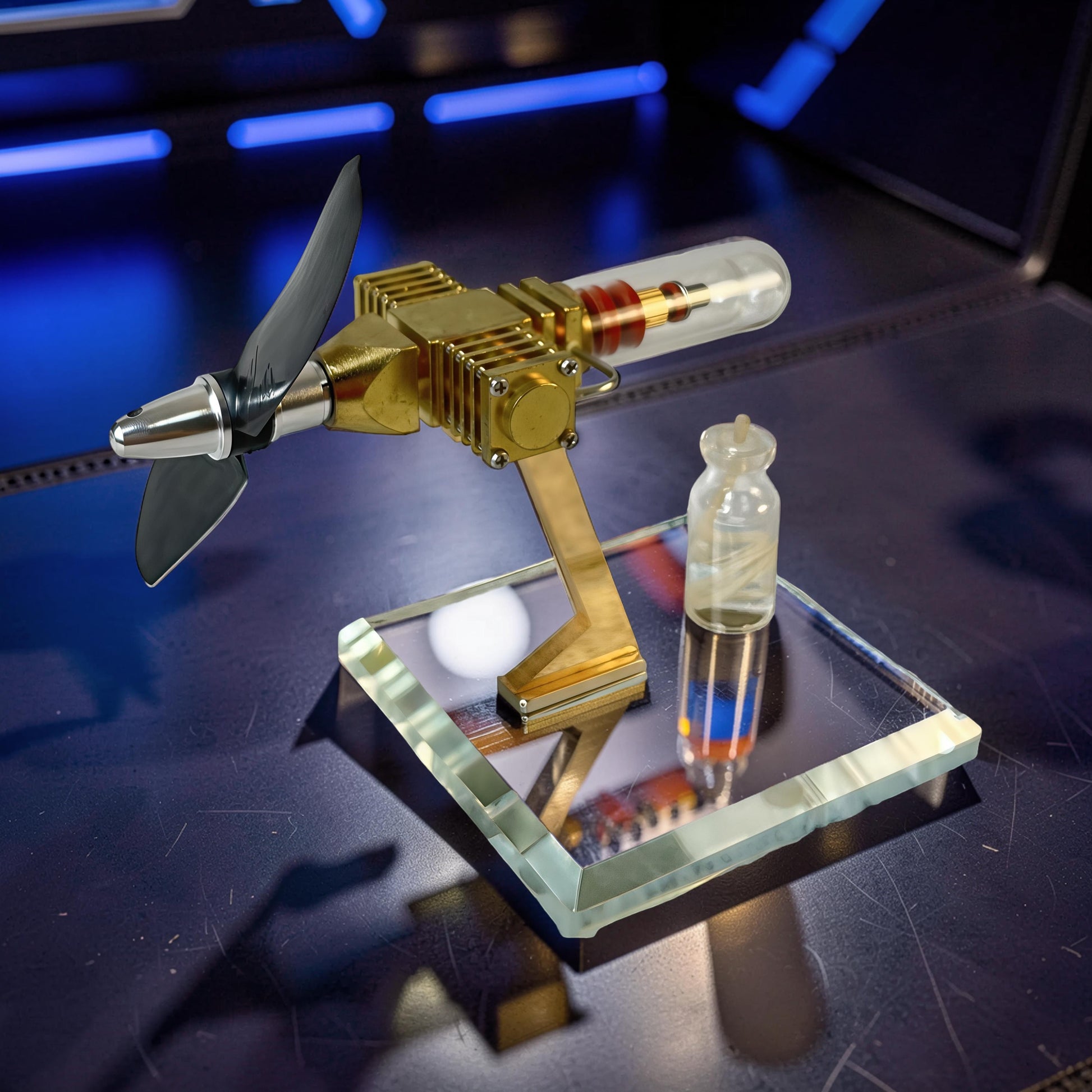 The Curious Mind Mini Aircraft Stirling Engine Display Model, a brass and metal engine with a propeller and glass cylinder, on a clear acrylic stand.
