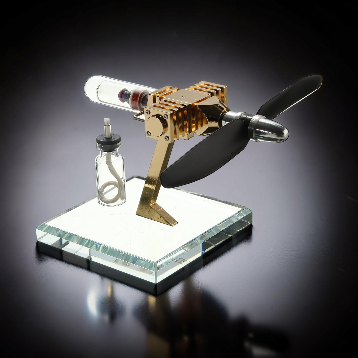 The Curious Mind Mini Aircraft Stirling Engine Display Model, gold metal, glass cylinder, black propeller, on a clear glass base.