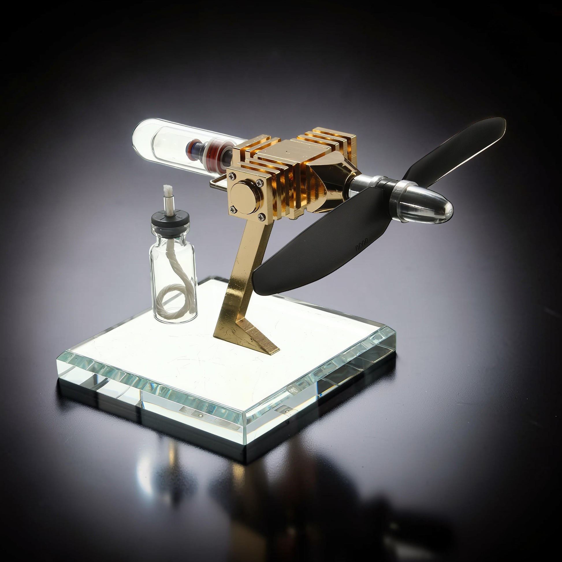 The Curious Mind Mini Aircraft Stirling Engine Display Model, gold metal, glass cylinder, black propeller, on a clear glass base.