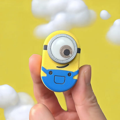 The Curious Mind Minion Pocket Push Pop Clicker fidget toy, yellow and blue metal and plastic, with one large eye and blue overalls.