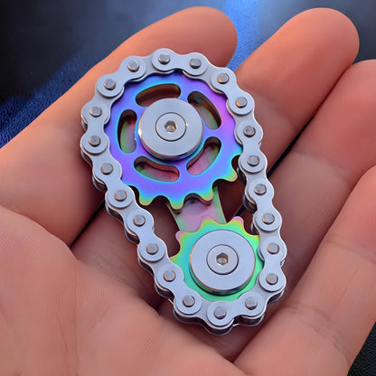 The Curious Mind Gear Chain Sprocket Spinner features a rainbow-colored metal gear set with a silver chain. This fidget toy is held in a hand.