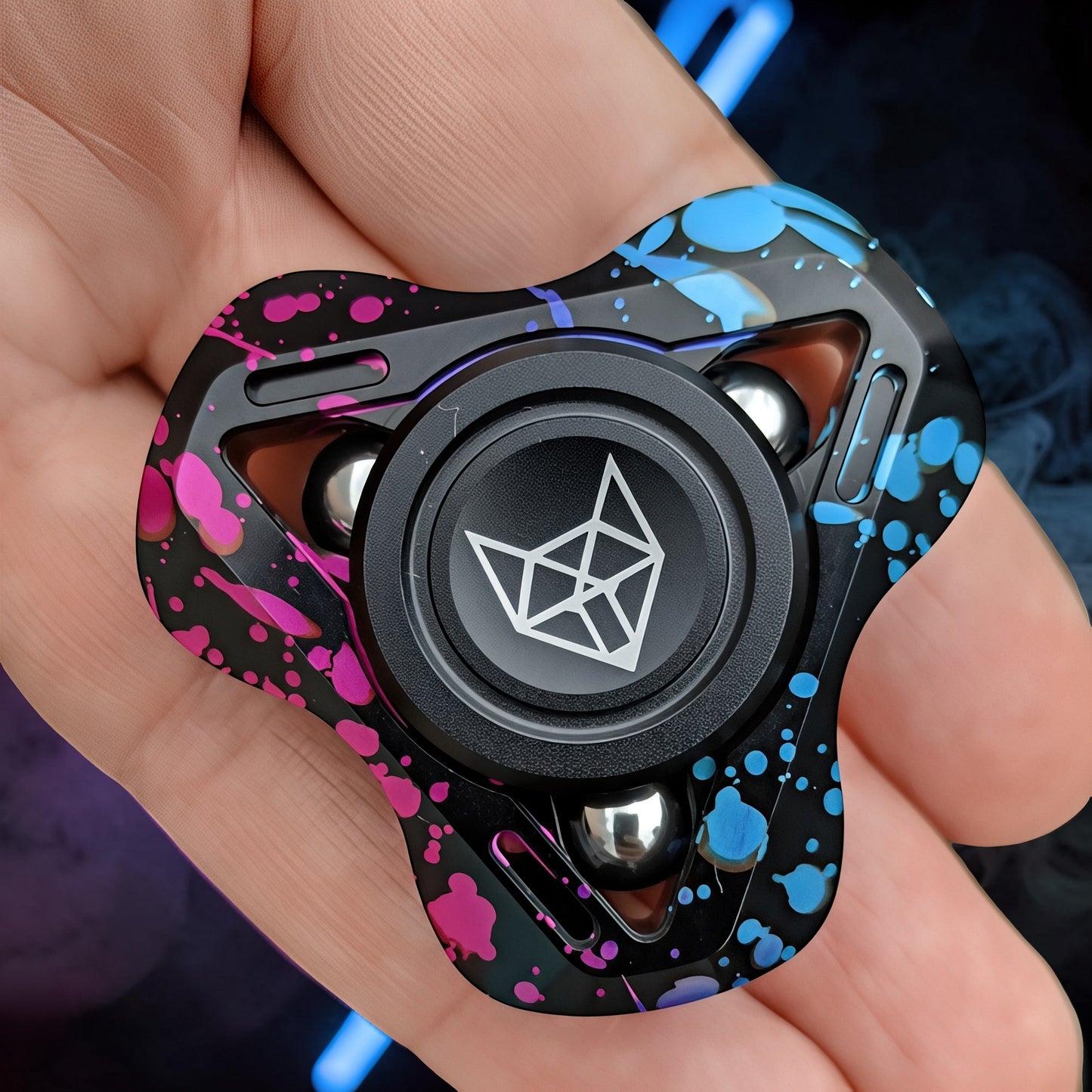 The Curious Mind Spirit Fox Glow Spinner with pink and blue paint splatter design on a black metal body, held in a hand.