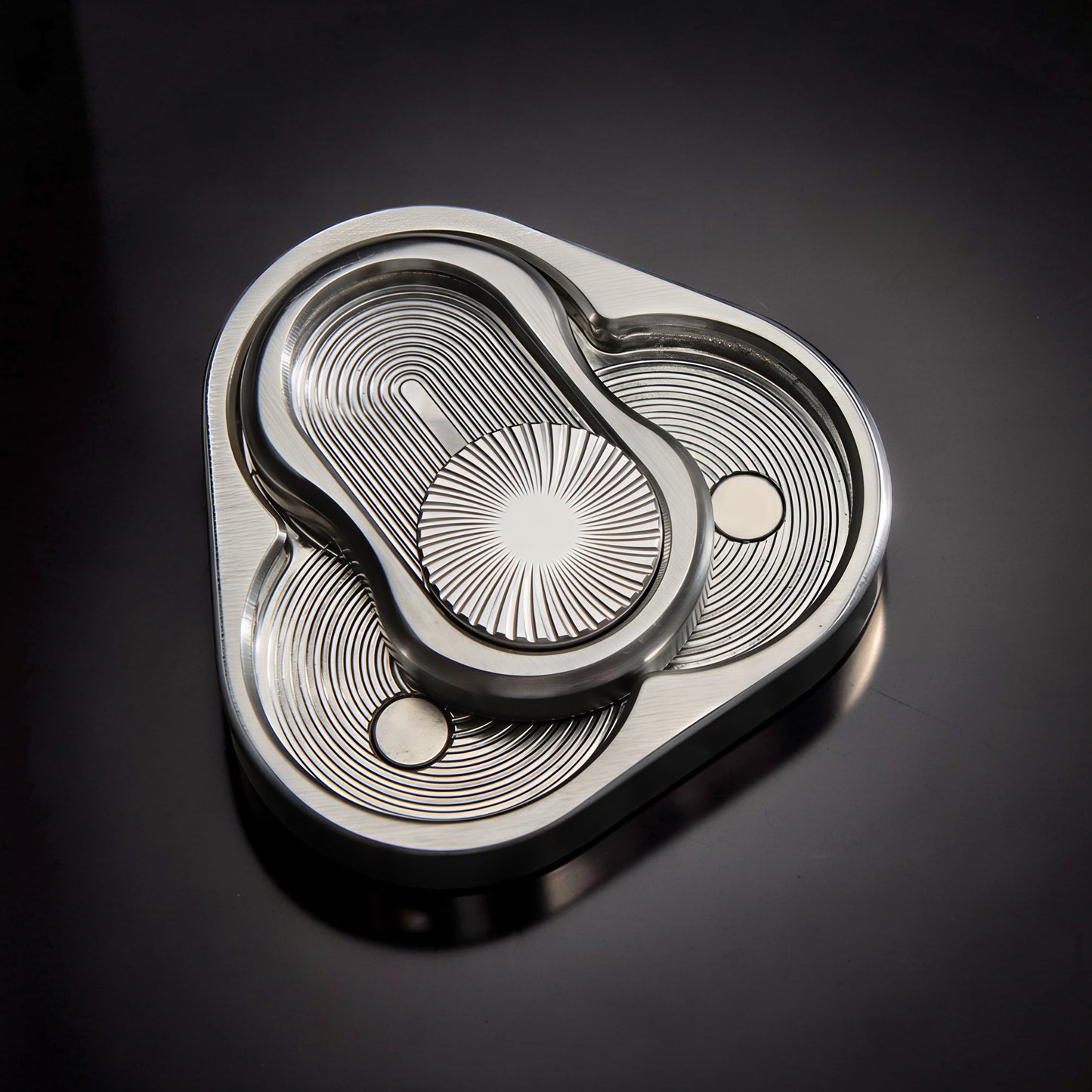 The Curious Mind Triangle Rotary Push Card fidget toy, made of brushed metal with intricate concentric circle patterns and a central sunburst design.